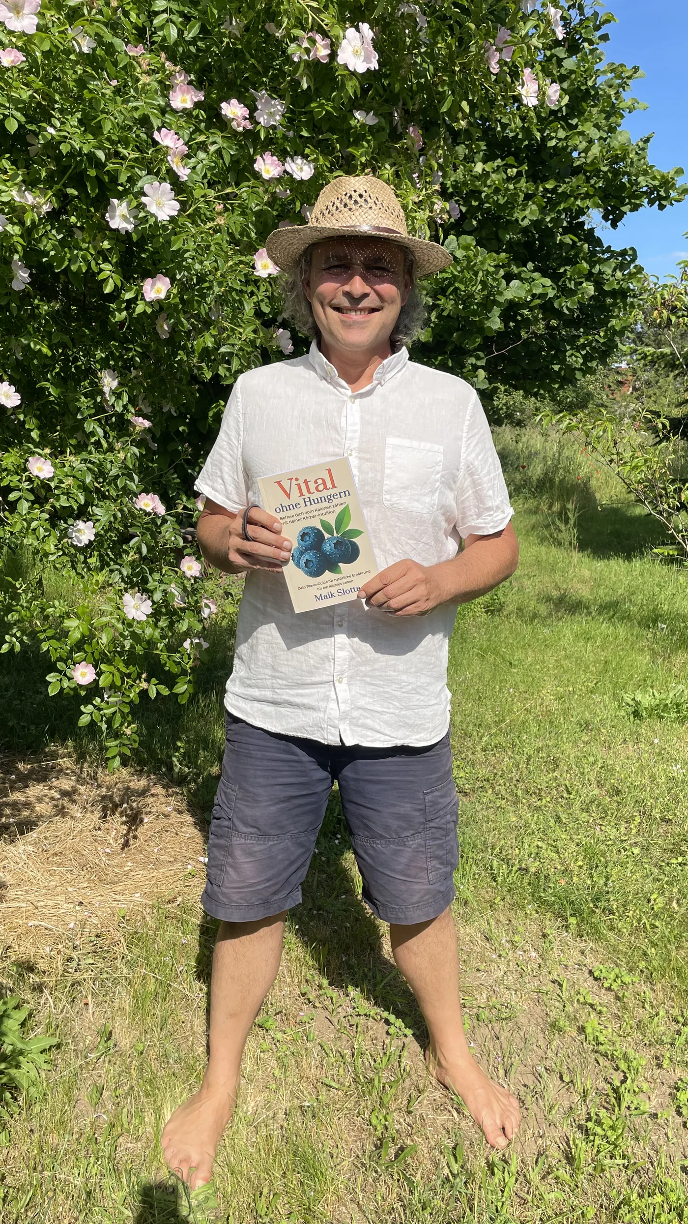 Maik steht barfuß auf einer Sommerwiese vor blühenden Wildrosen und hält sein Buch